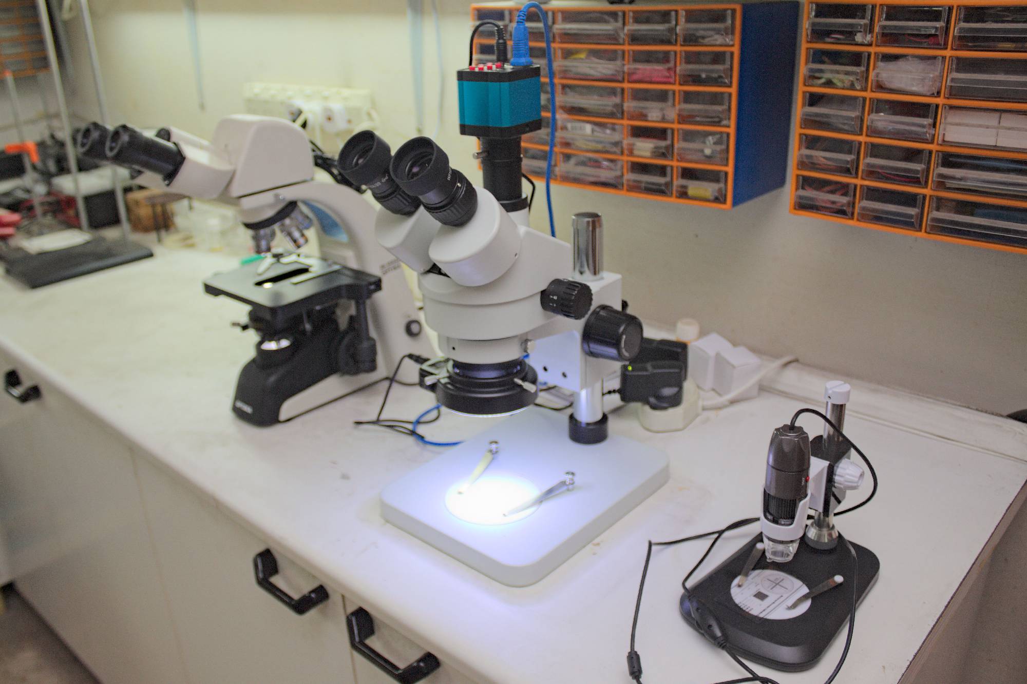 A high magnification binocular Optika light microcope, a stereomicroscope, and a USB operated microcope, placed upon an engineering laboratory work bench.